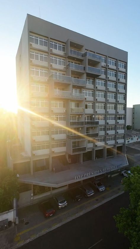 Prédio de apartamentos moderno e ensolarado ao pôr do sol, com árvores e torres vizinhas ao fundo.