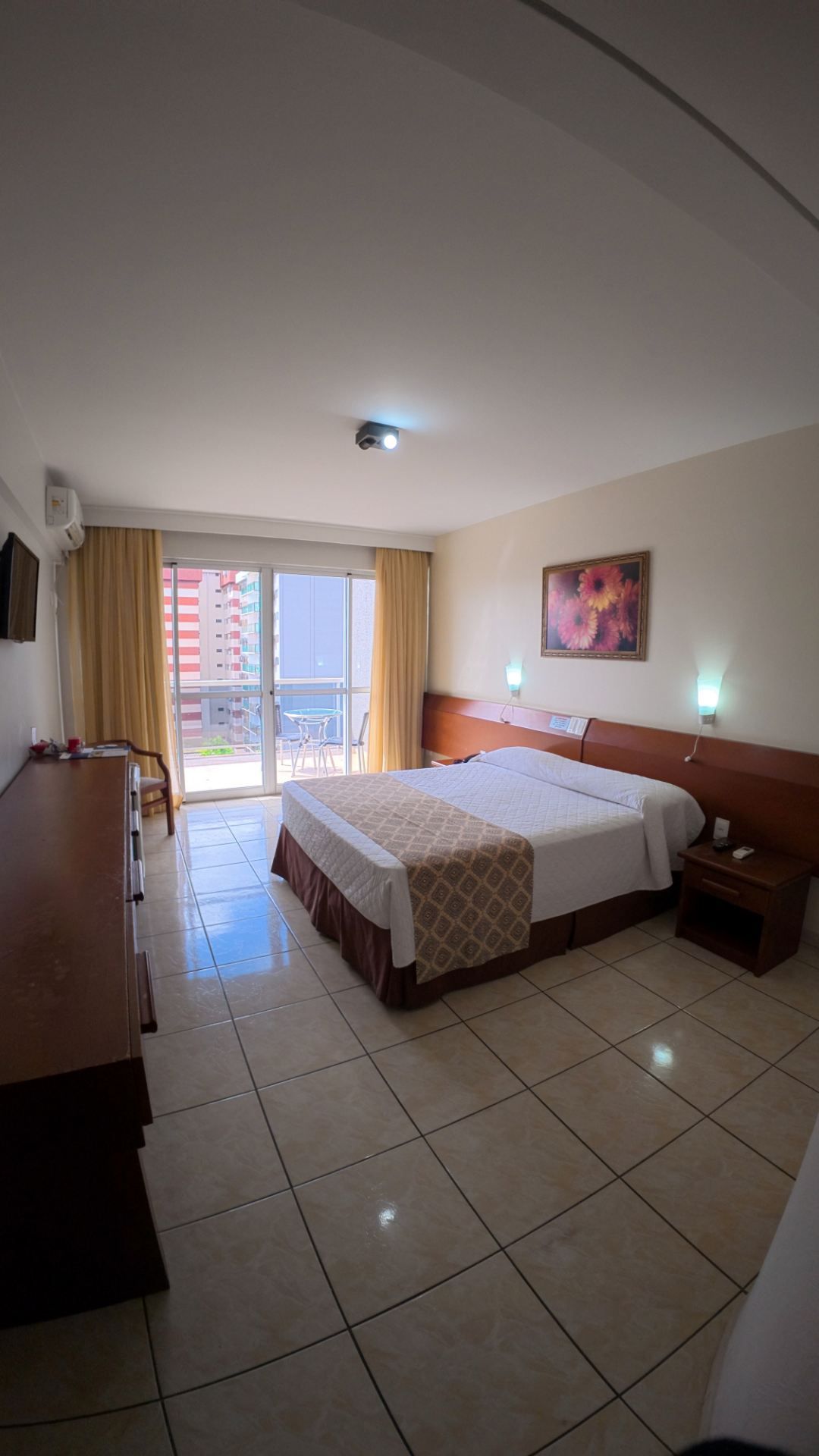 Quarto de hotel  com duas camas, piso de azulejo, escrivaninha, TV e portas de sacada que permitem a entrada de luz natural.