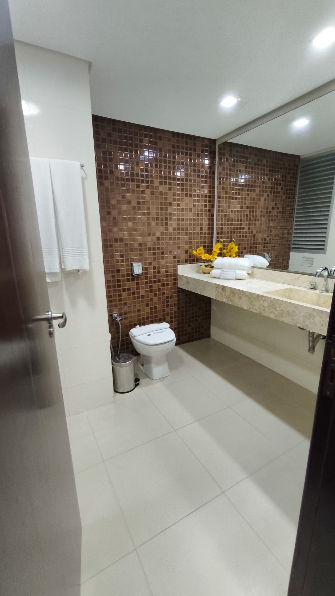 Banheiro moderno com azulejos bege, parede em mosaico marrom, pia branca, vaso sanitário e um grande espelho com pia.