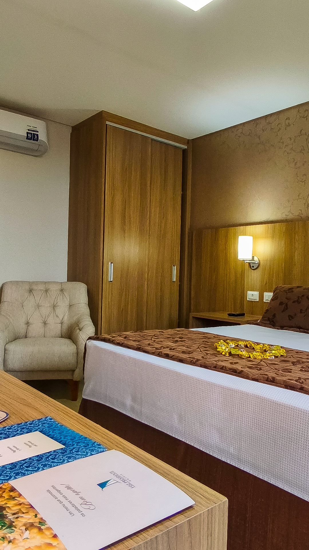 Quarto de hotel com cama, poltrona, guarda-roupa de madeira e luminária de parede ao lado de uma mesa de cabeceira.