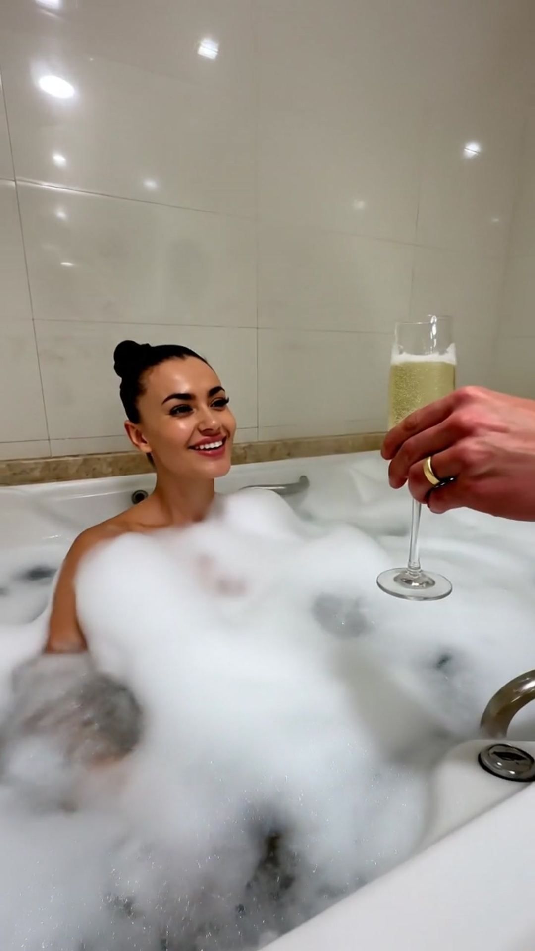 Pessoa relaxando em uma banheira de espuma enquanto outra mão lhe oferece uma taça de champanhe.