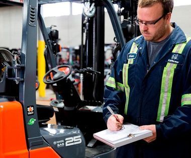 Un hombre está parado frente a una carretilla elevadora escribiendo en un portapapeles.