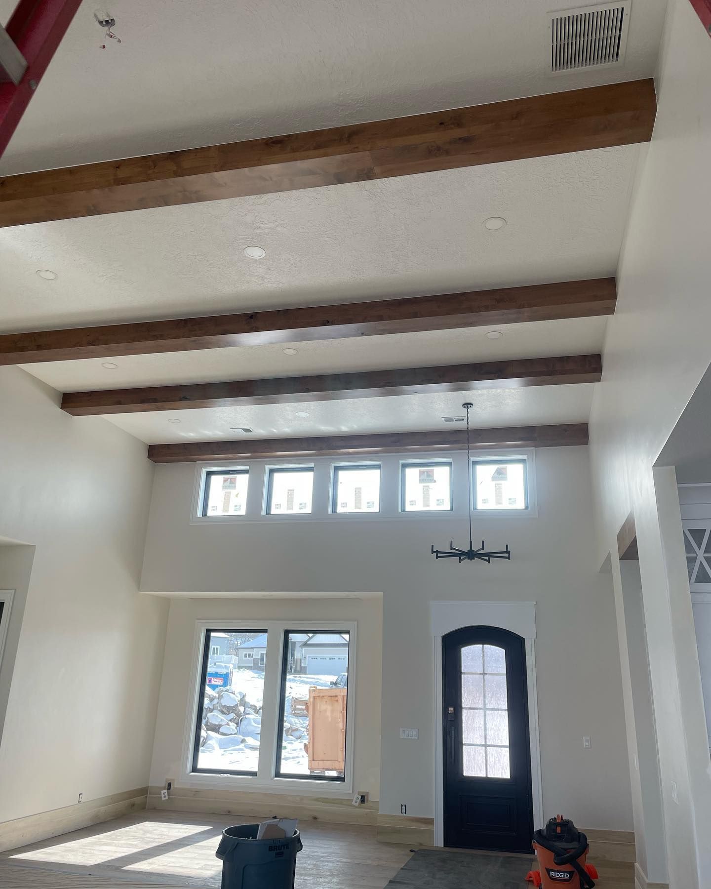 Interior of a house with high ceilings and wooden beams. Windows and a black door are visible.