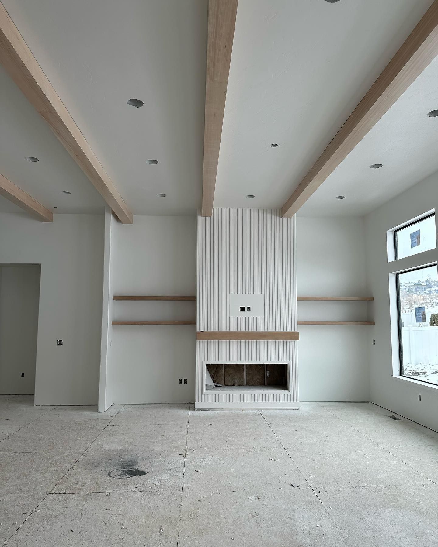 Unfinished interior of a room with a fireplace, shelves, and wood beams.  Gray floor, white walls and ceiling.