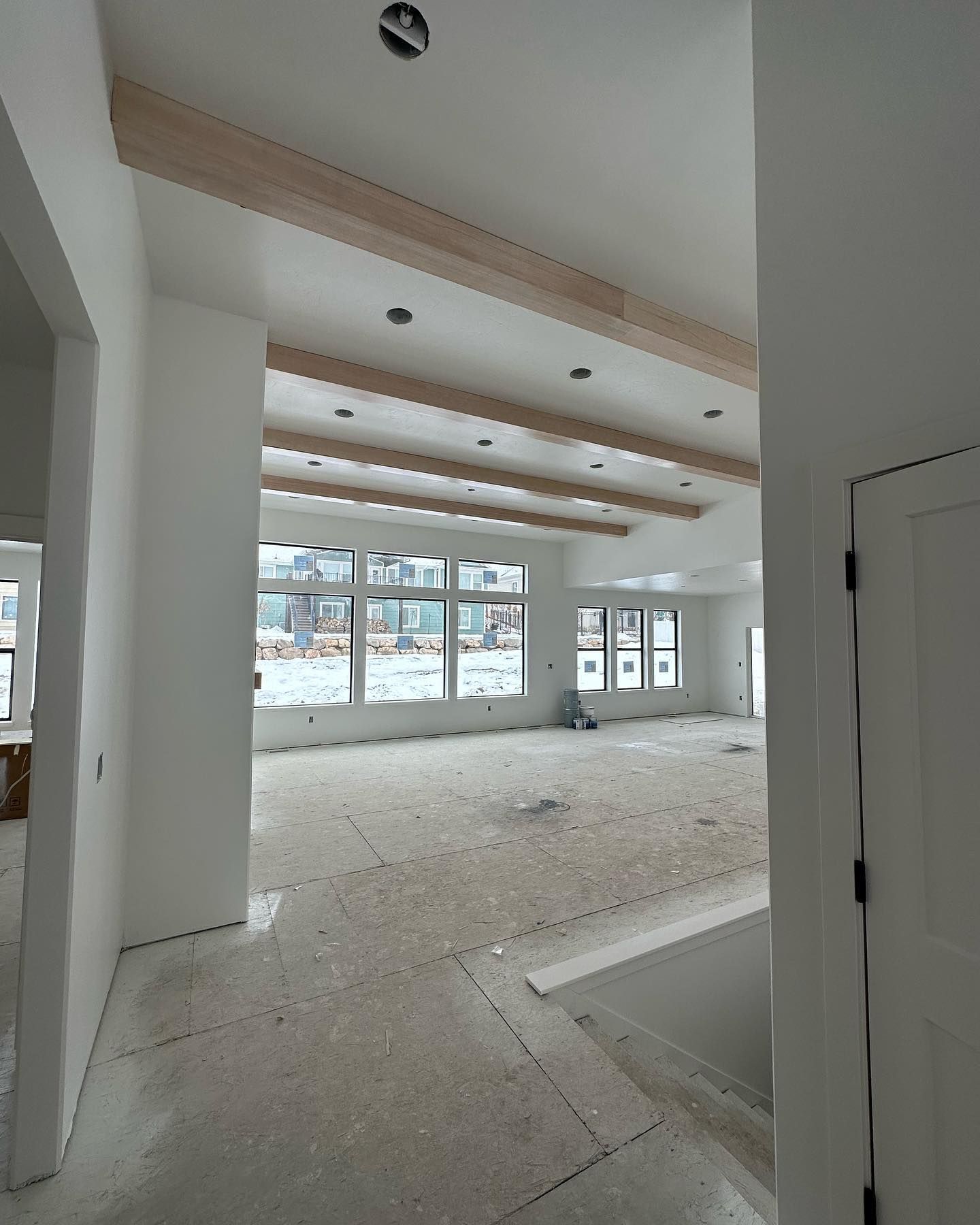 Interior of a home under construction. White walls and ceiling, light wood beams, large windows, and unfinished floor.