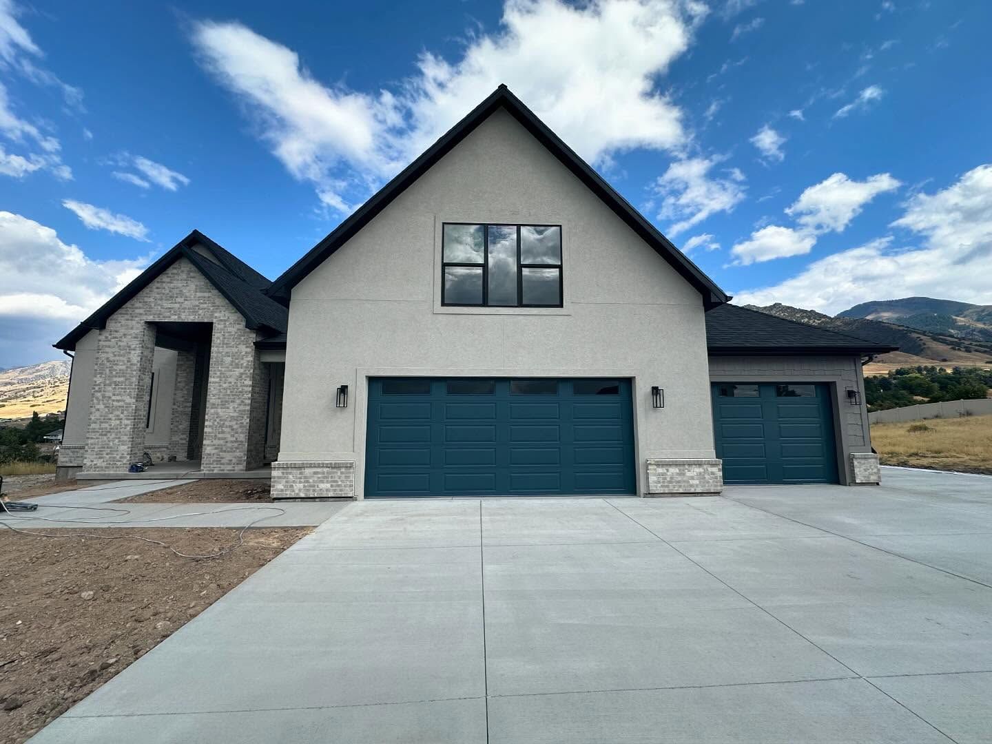 Modern house with blue-green garage doors, stone accents, and a long concrete driveway under a blue sky.