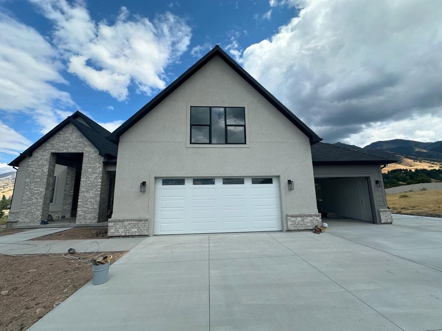 Front of a modern house with gray stucco, black roof, and garage. Overcast sky, long driveway.