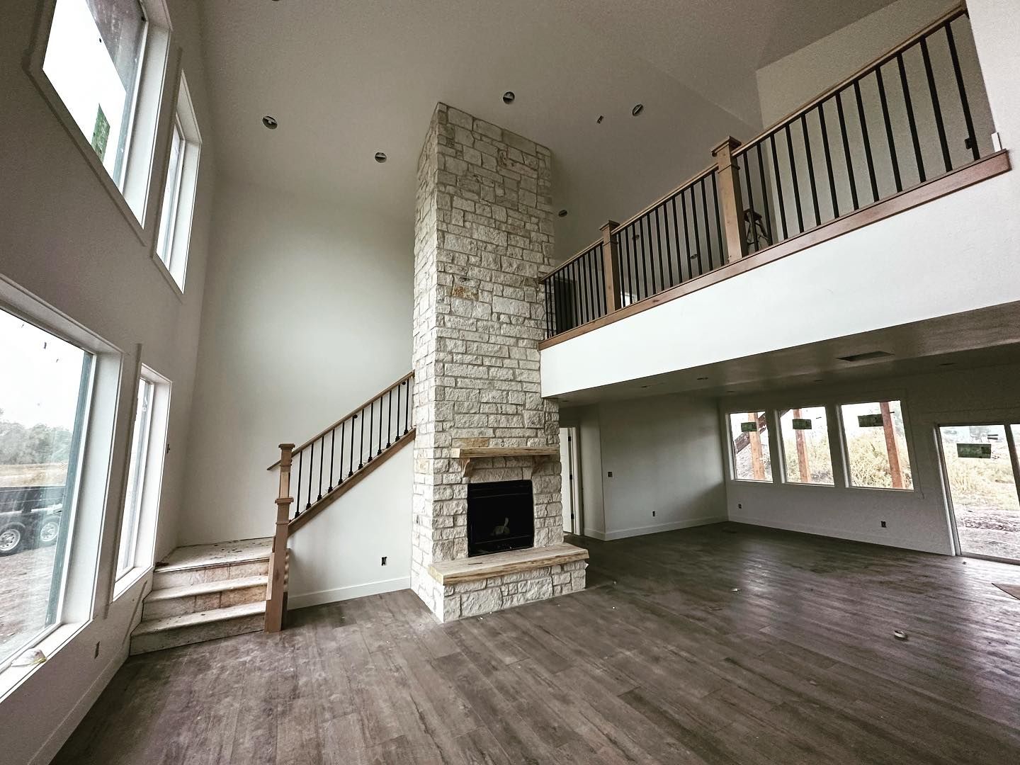 Spacious interior with stone fireplace, wooden staircase, and open second-floor balcony. Large windows and hardwood floor.