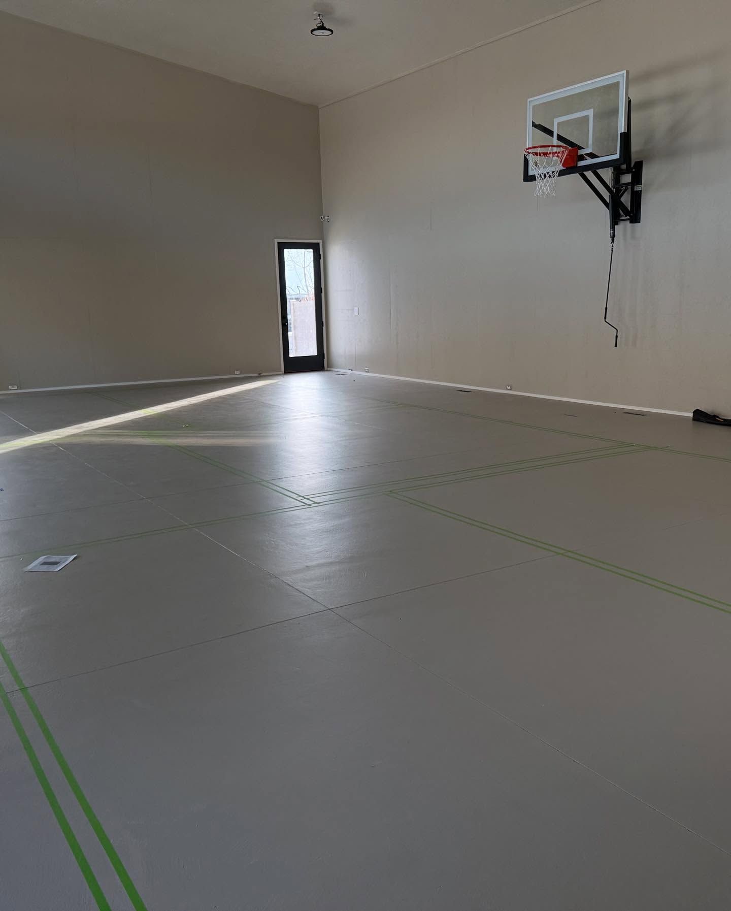 Empty indoor gym with light-colored flooring, basketball hoop, and a door in the back.
