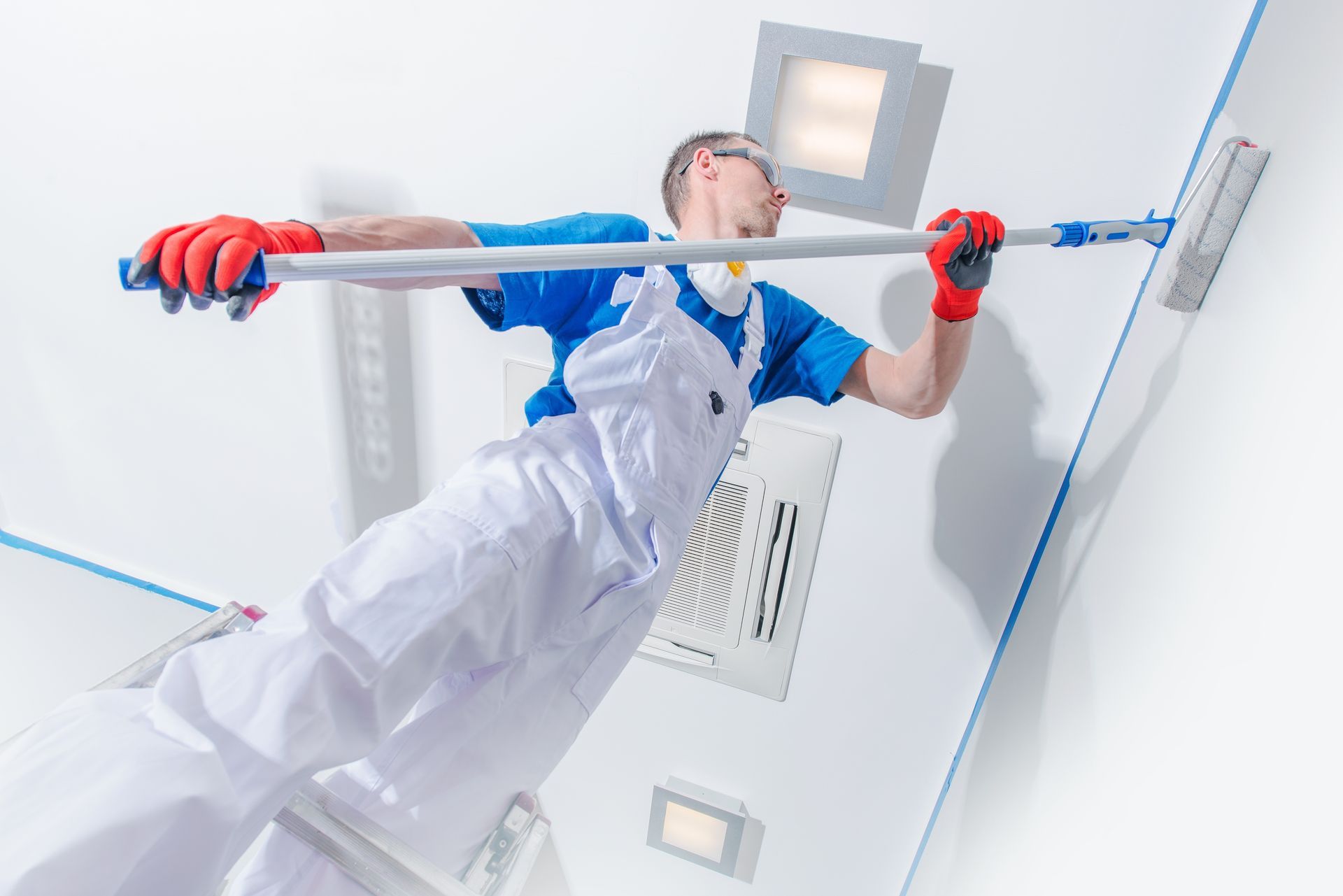 Painter in blue shirt and white overalls uses a roller on a wall, standing on a ladder.