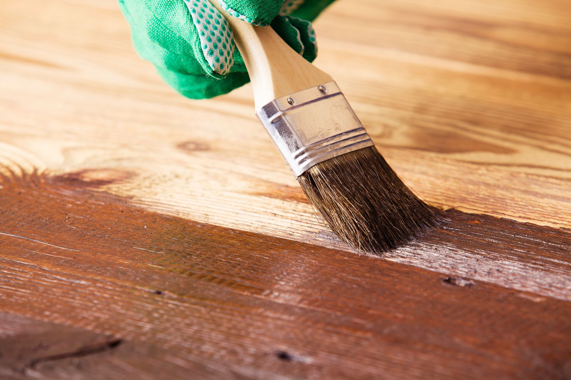 Gloved hand using a brush to apply brown stain to wooden planks.
