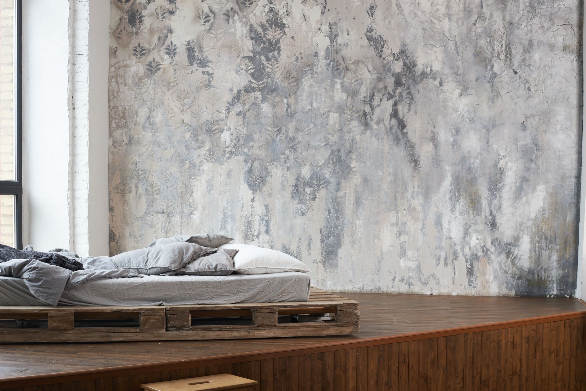 Bed with rumpled gray bedding on a wooden pallet, against a textured gray wall, beside a window.