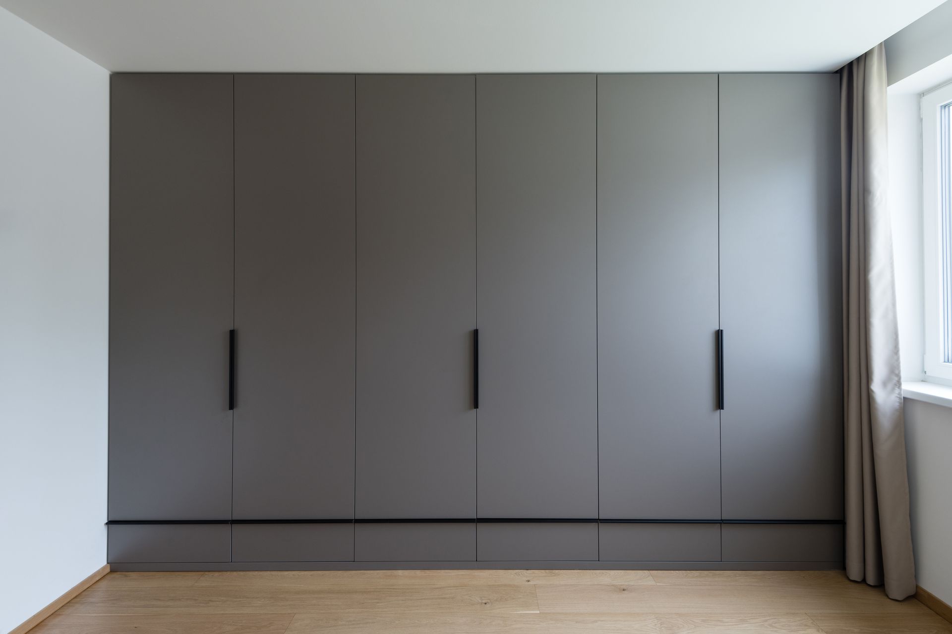 Gray built-in closet with black handles in a room with light wood floors and a window with a curtain.