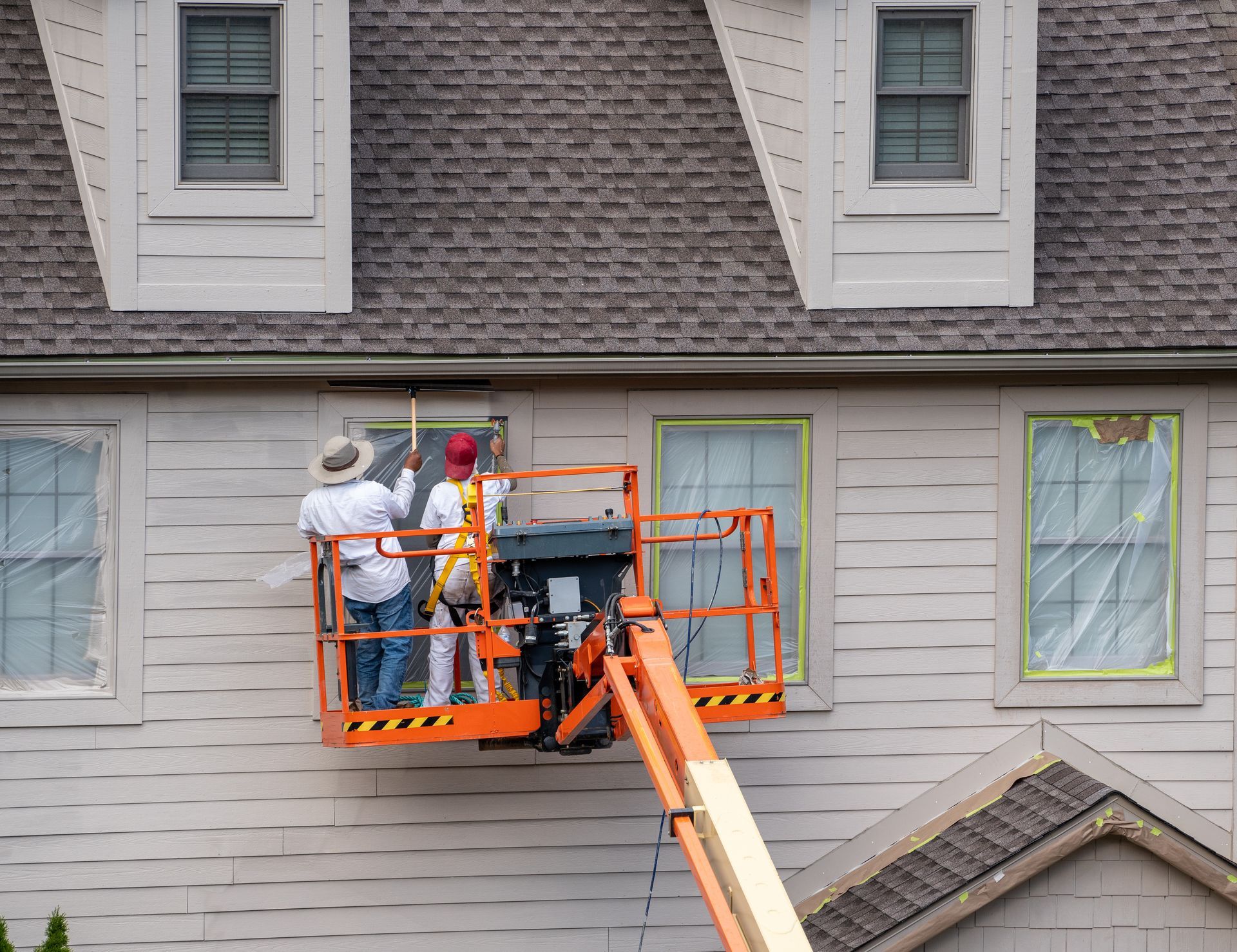 Two workers painting a house from a lift platform. One paints, the other assists.