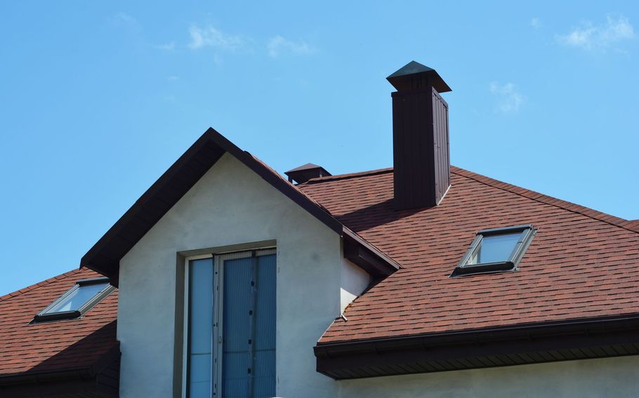 A brown shingled roof with a chimney, a dormer window, and two skylights against a clear blue sky.
