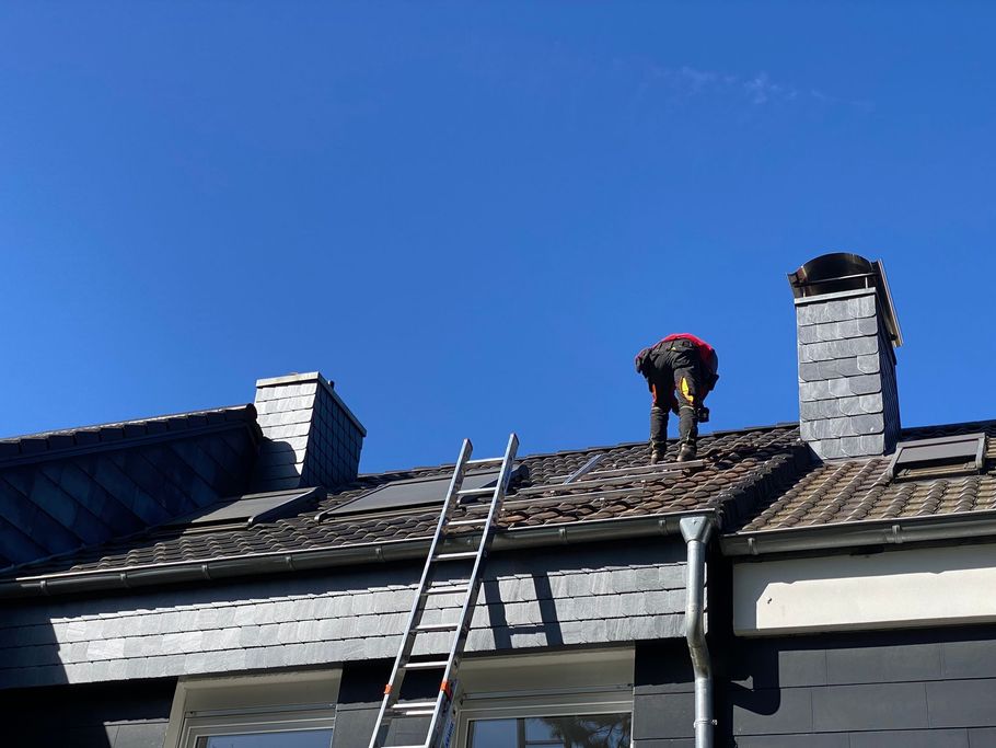 A person in work clothes stands on a tiled roof near a chimney, with a ladder leaning against the side of the house.