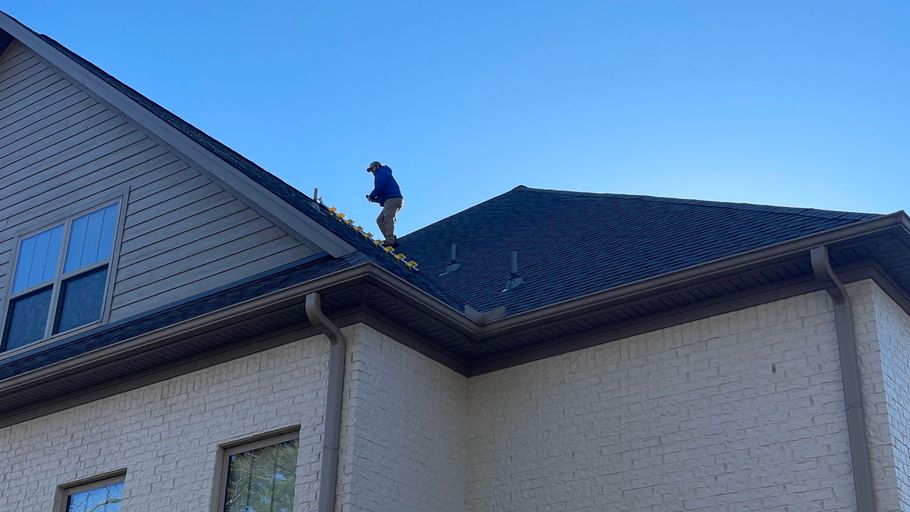 A person wearing a blue shirt works on the shingled roof of a two-story beige brick house under a clear blue sky.