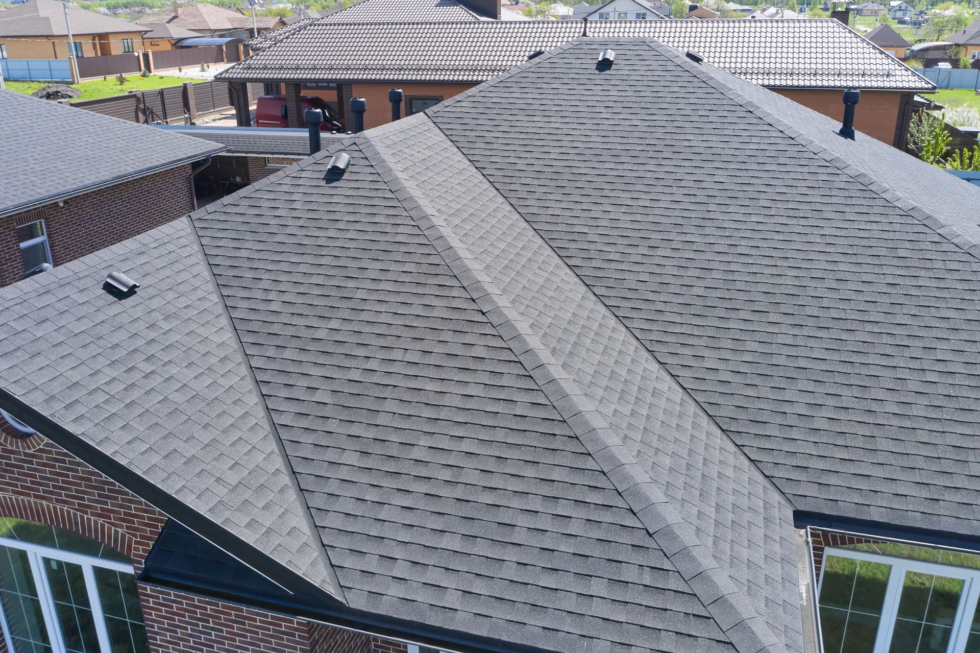 A high-angle view of a grey shingled roof on a brick house with multiple pitches, valleys, and vent stacks.