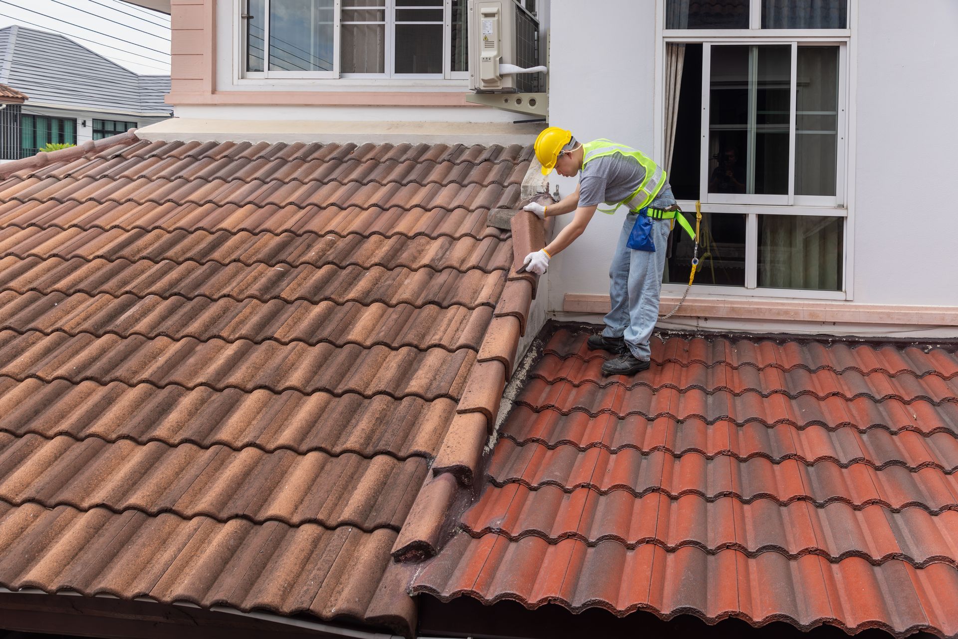 A construction worker in a yellow hard hat and safety harness repairs terracotta roof tiles on a residential building.