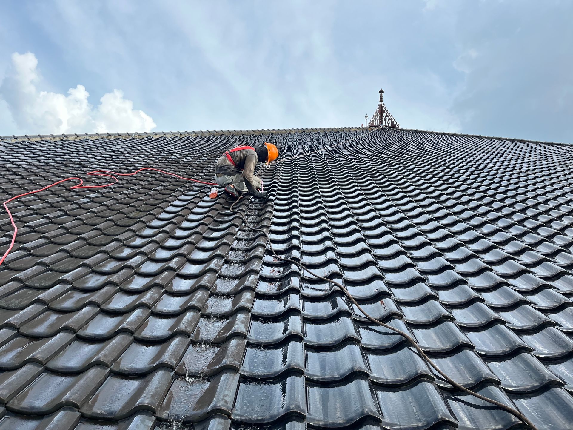 A worker in a high-visibility orange hard hat performs maintenance on a dark-tiled roof under a cloudy sky.