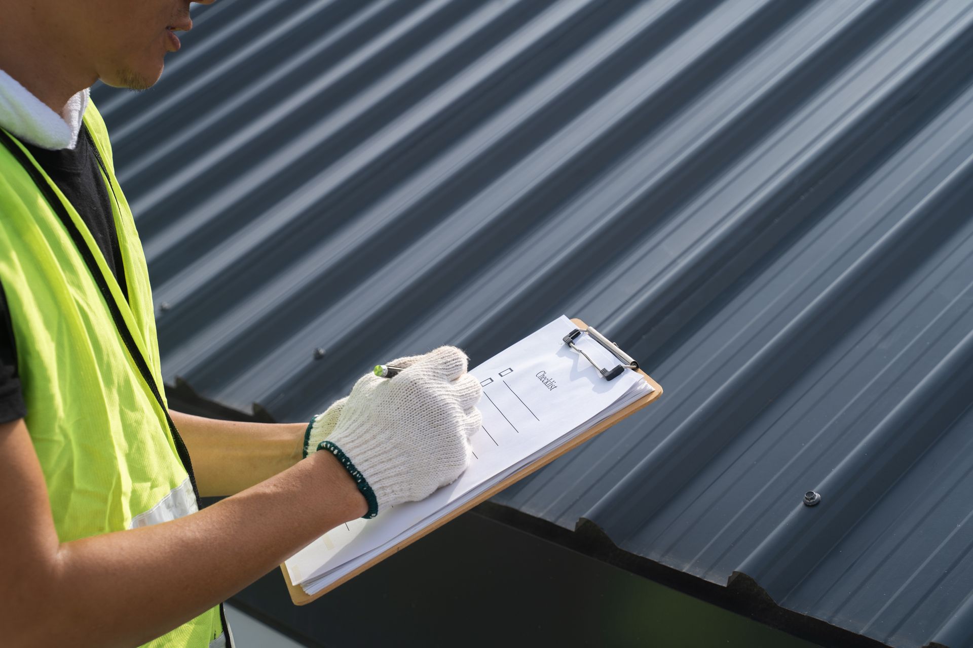 A worker in a high-visibility vest and work gloves writes on a clipboard while inspecting a dark metal roof.