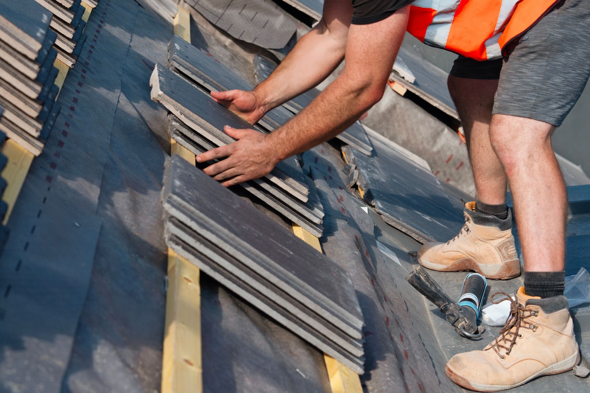 A worker in a high-visibility vest installs dark roof tiles onto wooden battens on a sloped roof.