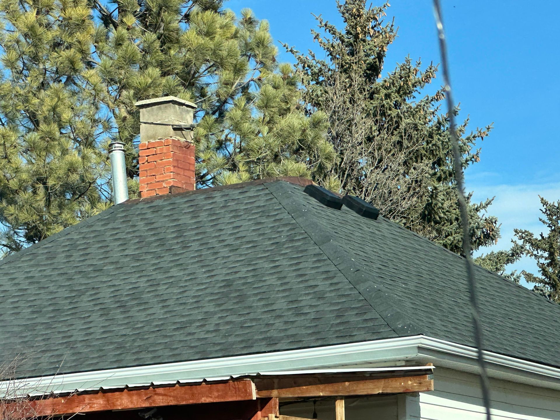 A brick chimney and a metal vent pipe stand on a dark shingled hip roof, set against a background of evergreen trees.