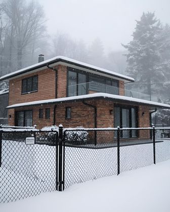 Two-story brick house with large windows, covered in snow, behind a chain-link fence.
