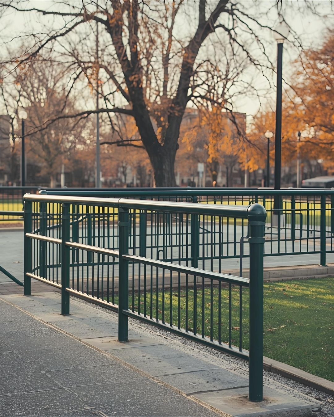 Green metal fence bordering a paved pathway and grassy area with a large tree in the background.