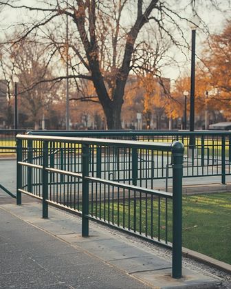 Green metal fence bordering a paved pathway and grassy area with a large tree in the background.