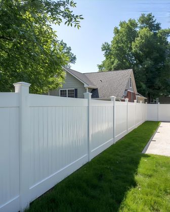 White vinyl fence in a grassy yard, partially obscuring a house and trees under a blue sky.