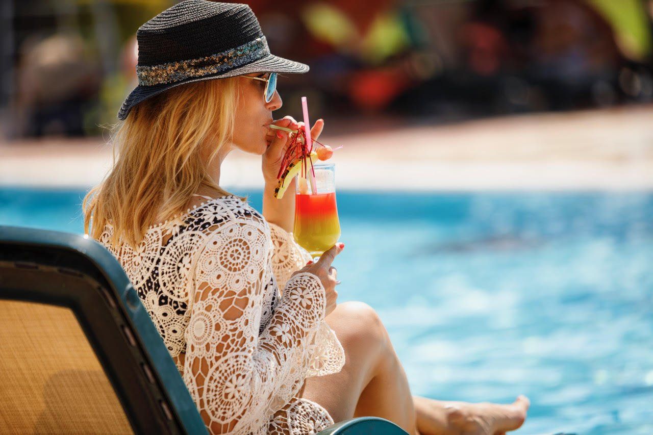 A woman is sitting in a chair by a swimming pool drinking a cocktail.