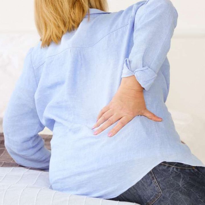 Woman in blue shirt touches lower back, possibly in pain, sitting on a bed.