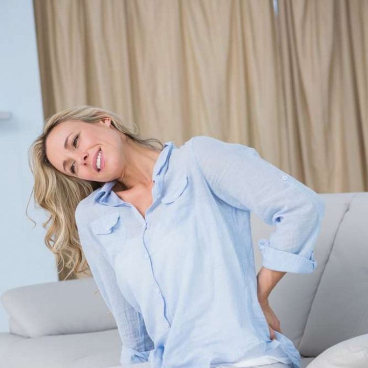 Woman with back pain, leaning on a sofa, grimacing in a light-filled room.