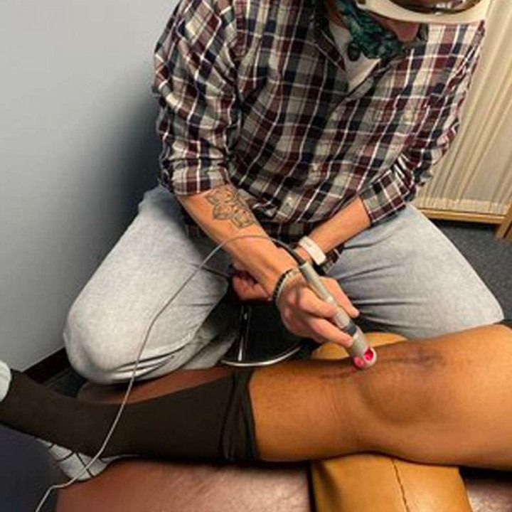 A person using a handheld device on a patient's knee. The device has a red light. Indoors.