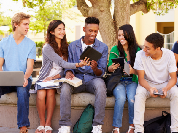 Students sitting outdoors on a campus bench, chatting and studying with laptops and notebooks.