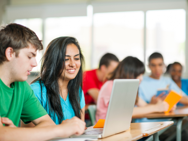 Students collaborate around a laptop in a bright classroom, smiling and working together.