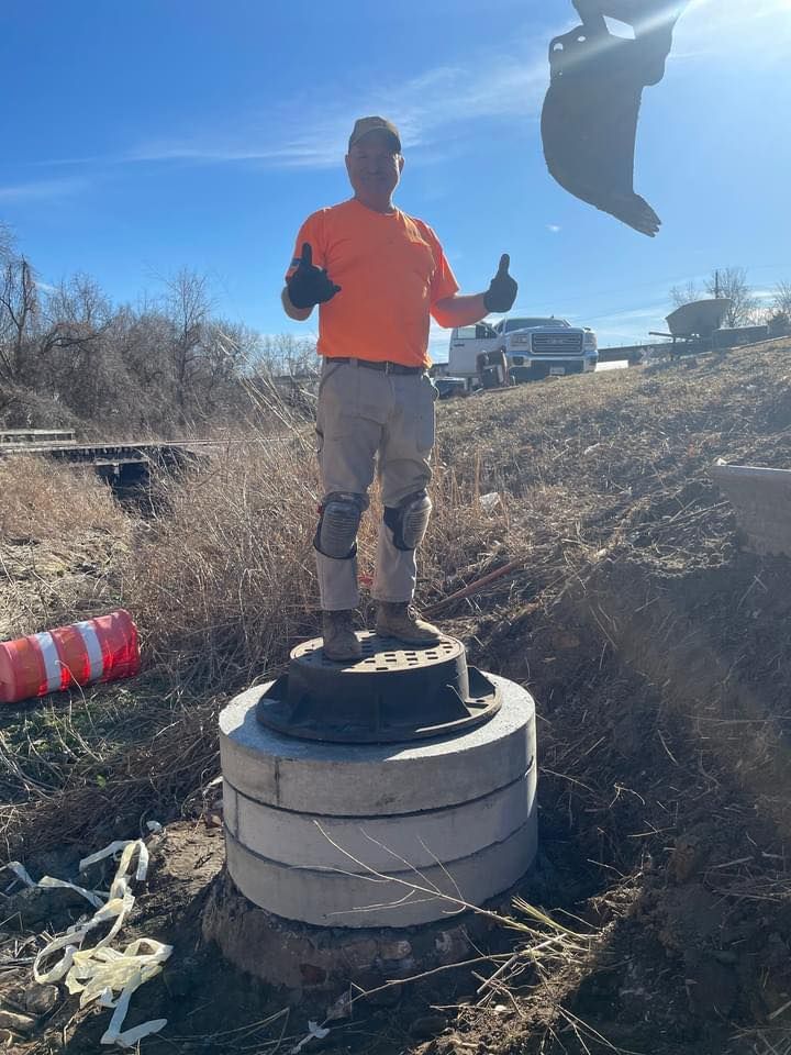 Construction worker stands on concrete rings, giving thumbs-up. Bright orange shirt, beige pants, blue sky.