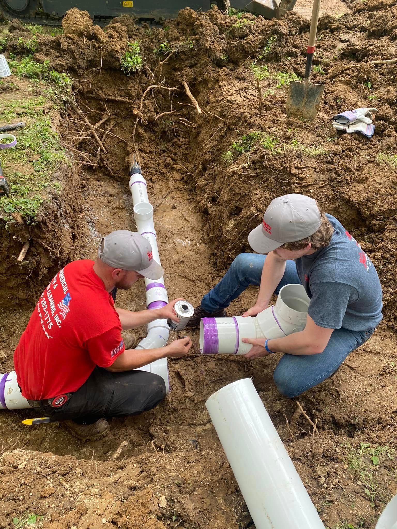 Two men installing PVC pipes in a trench outdoors. One holds pipe, the other fits parts together.