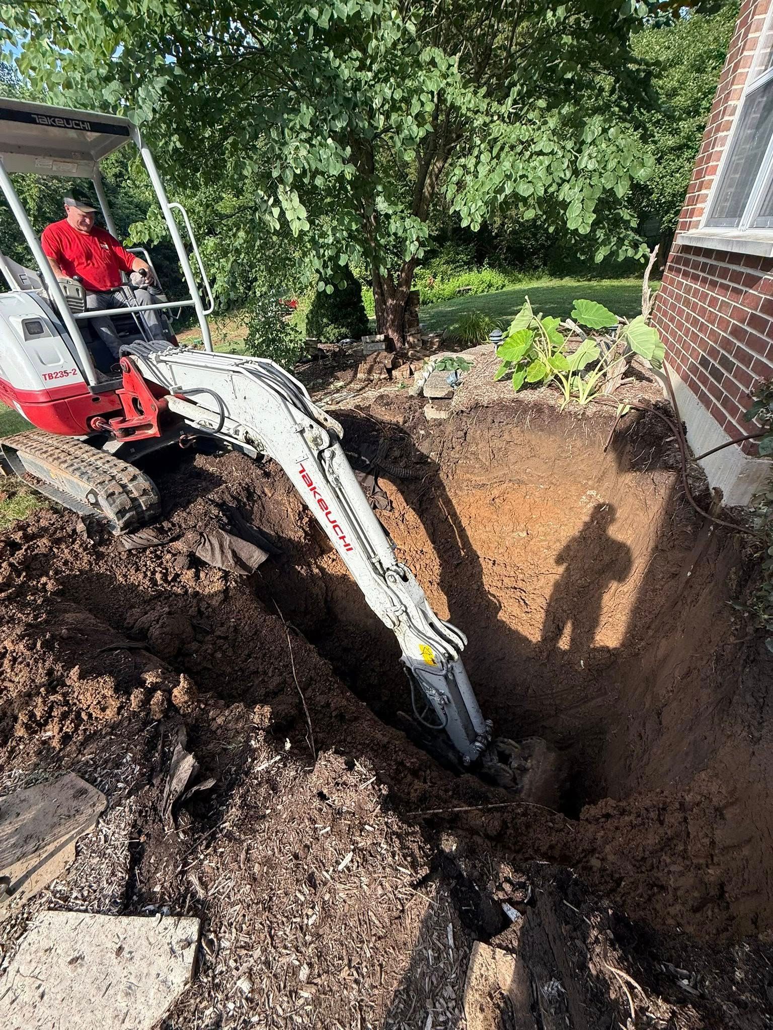 A small excavator digs near a house, operated by a person in a red shirt. Soil is excavated and piled.