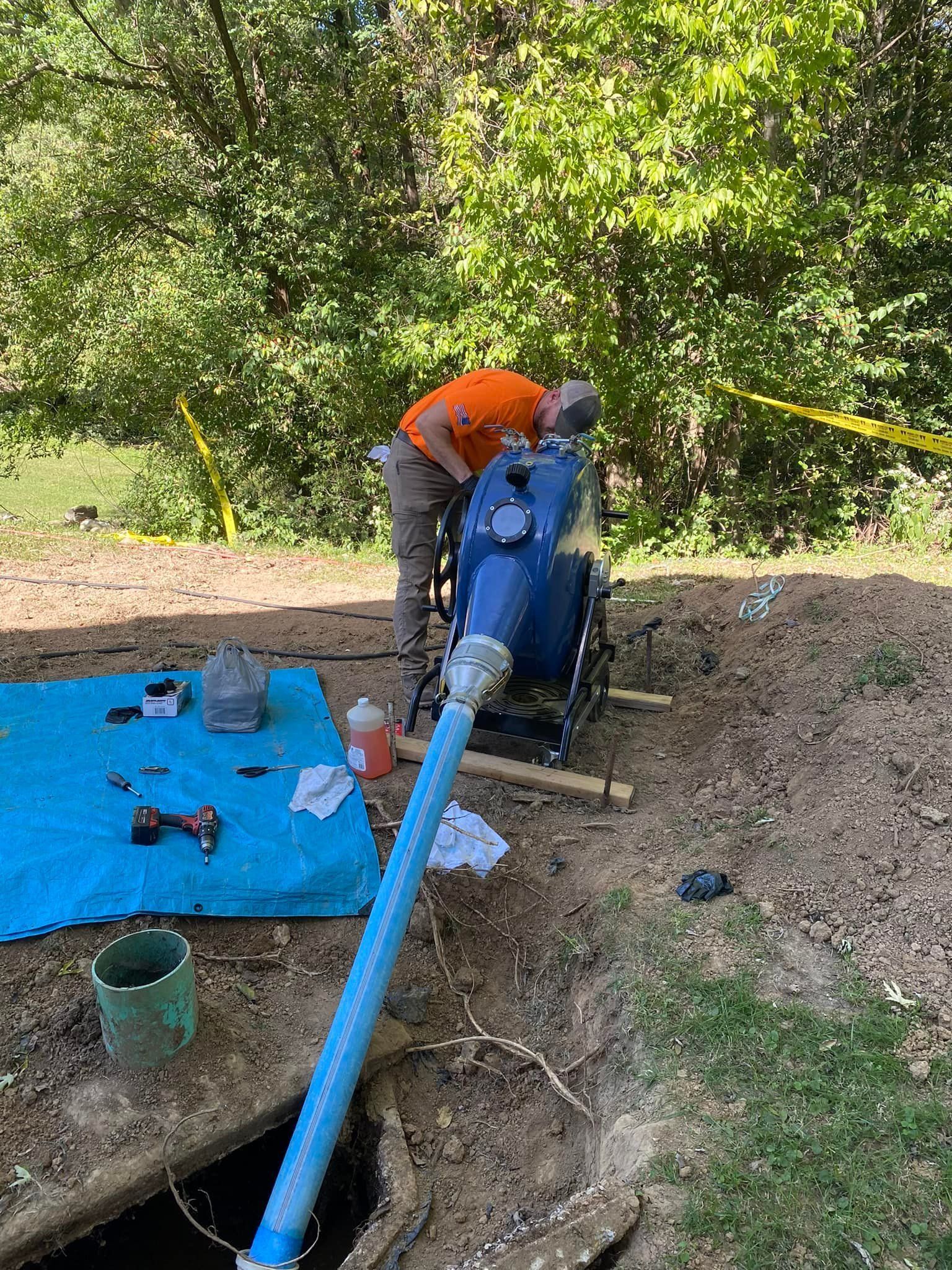 Man working on blue tank with a hose outdoors near a hole and foliage.
