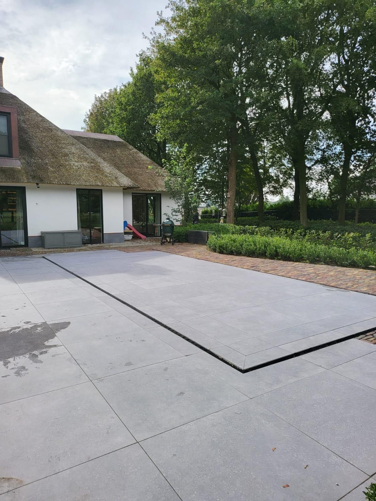 Exterieur van een huis met een rieten dak, een betonnen terras en een tuin met bomen.
