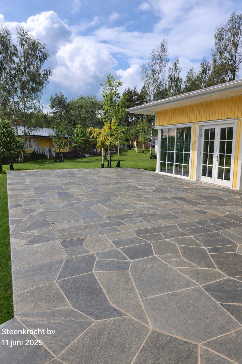 Terras met donkergrijze natuurstenen tegels op de MagicPool, een bakstenen muur, tuinmeubilair en een huis op de achtergrond.