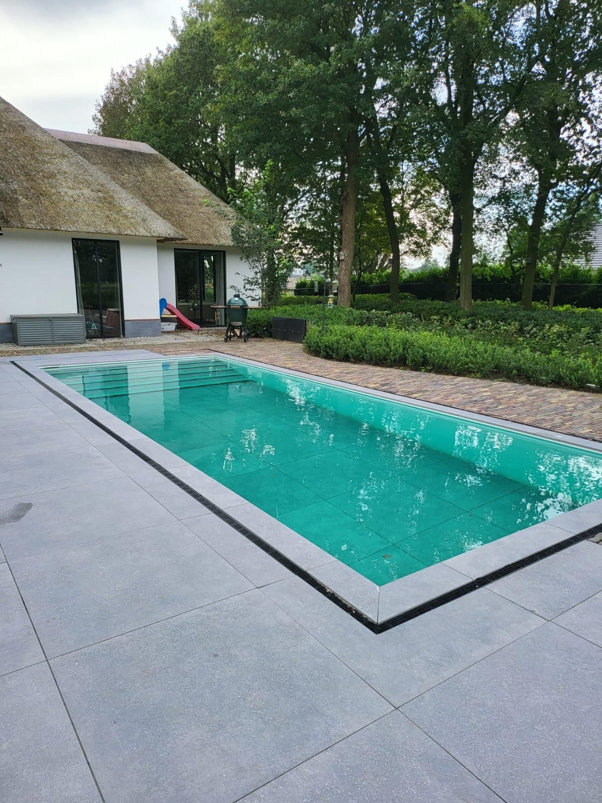 Rechthoekig zwembad op een grijs stenen terras naast een wit gebouw met een rieten dak. Groen water.