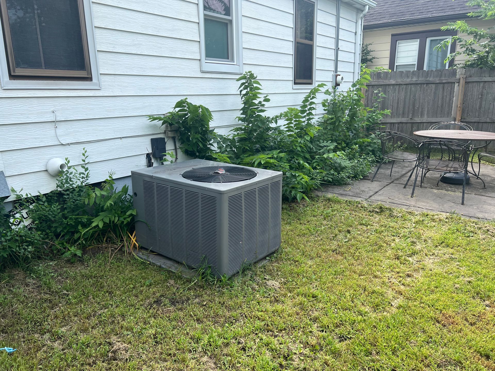 Outdoor AC unit beside a white house, with grass, shrubs, and a patio table nearby