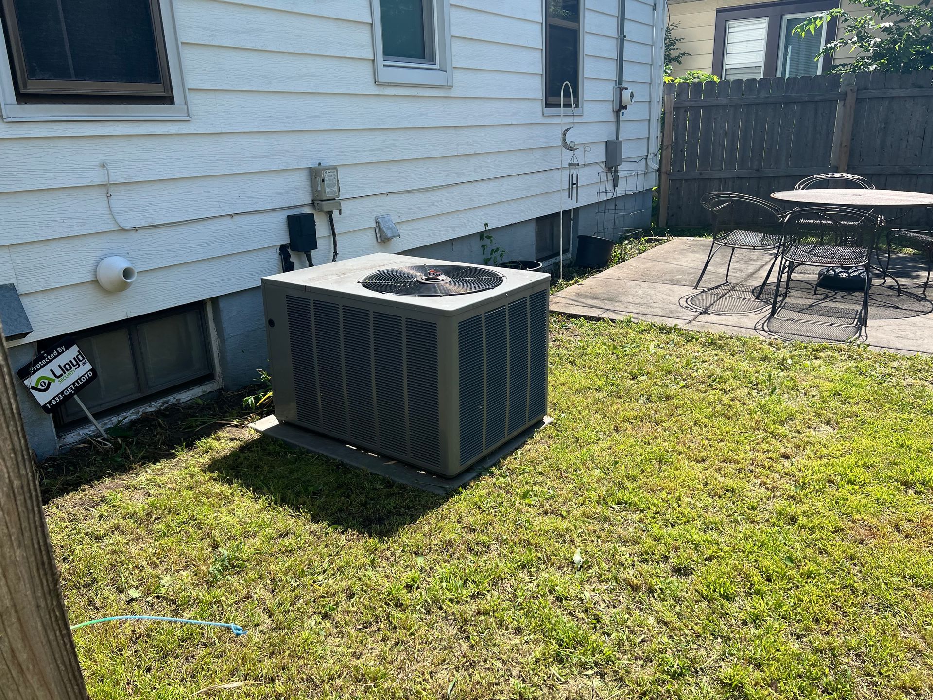 Outdoor HVAC unit beside a white house on a grassy lawn, with a patio and fence in the background