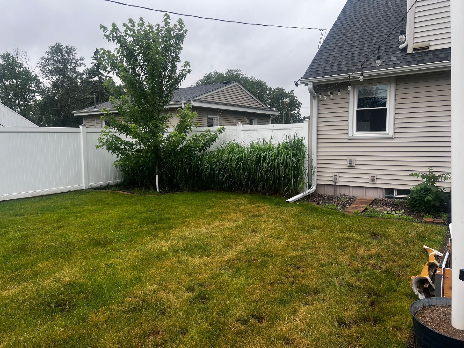 Backyard with green lawn, white fence, small tree, and beige house under a cloudy sky