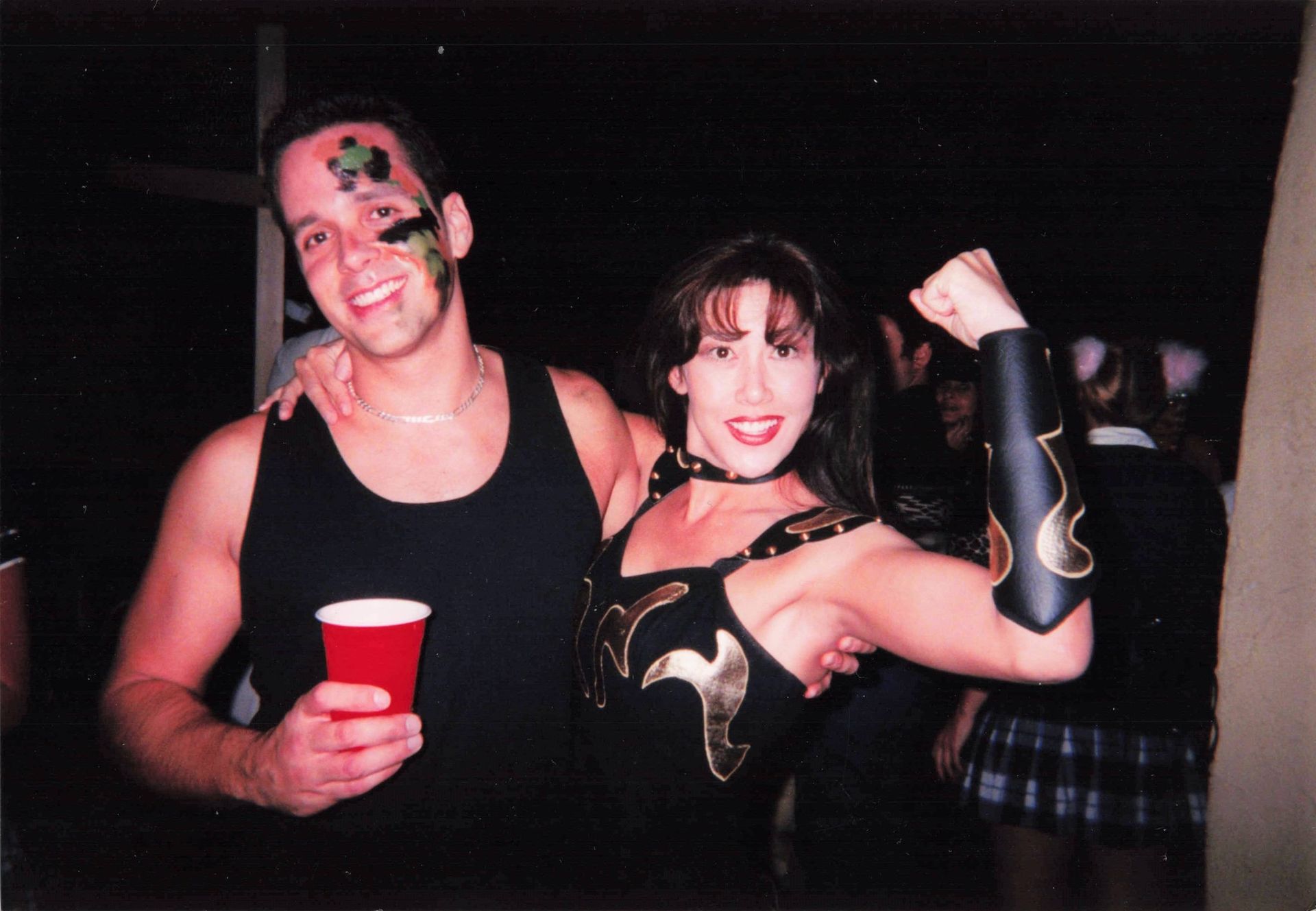 A man in a black tank top holds a red cup next to a woman in a black and gold costume