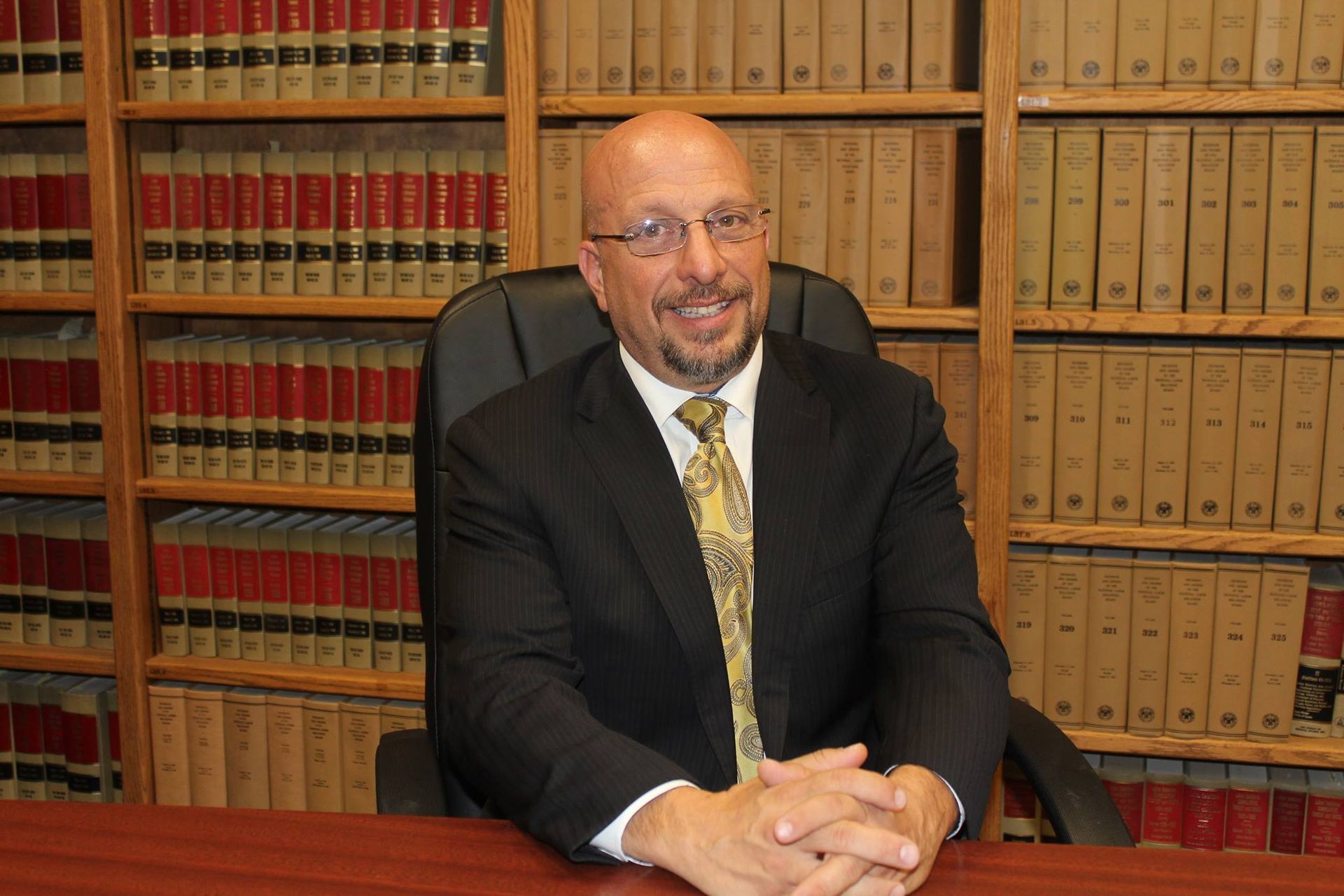 A man in a suit and tie is sitting at a desk in front of a bookshelf.