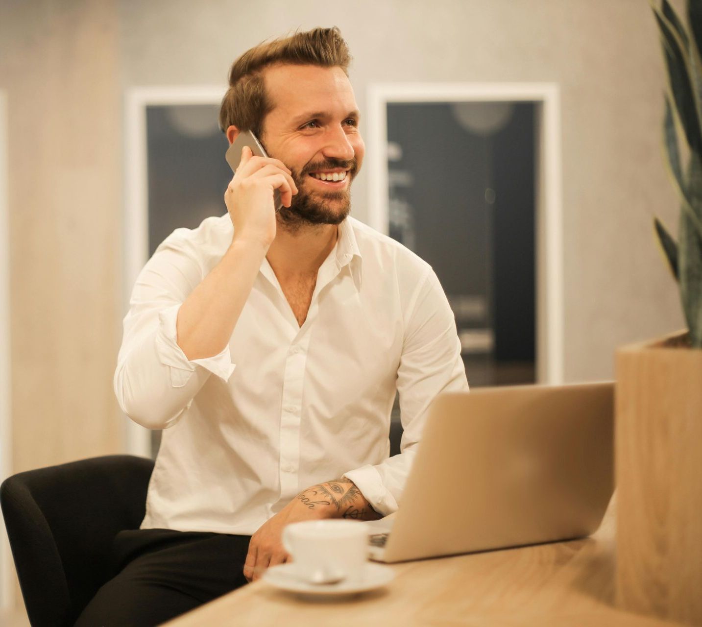 A man is sitting at a desk with a laptop and talking on a cell phone.